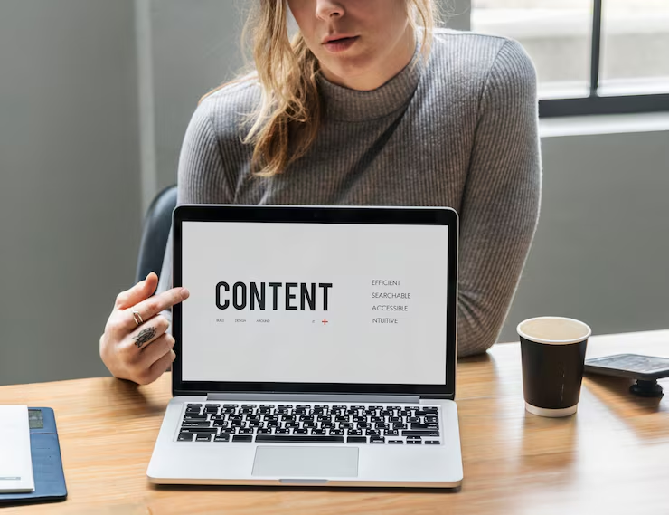 Person seated at a table holding open a laptop displaying the word “CONTENT,” with a coffee cup beside it.